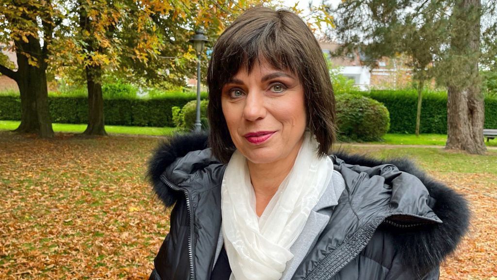 Portrait of Victoria, who is looking at the camera with a small smile. She has straight, dark, bobbed hair with a fringe. She is wearing a black puffa jacket with fur round the hood and a light white scarf. She is pictured in a park, with trees and grass behind her and leaves on the ground.