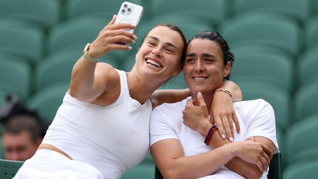 Aryna Sabalenka has her right arm outstretched holding a phone as she takes a selfie with Ons Jabeur on the side of a tennis court