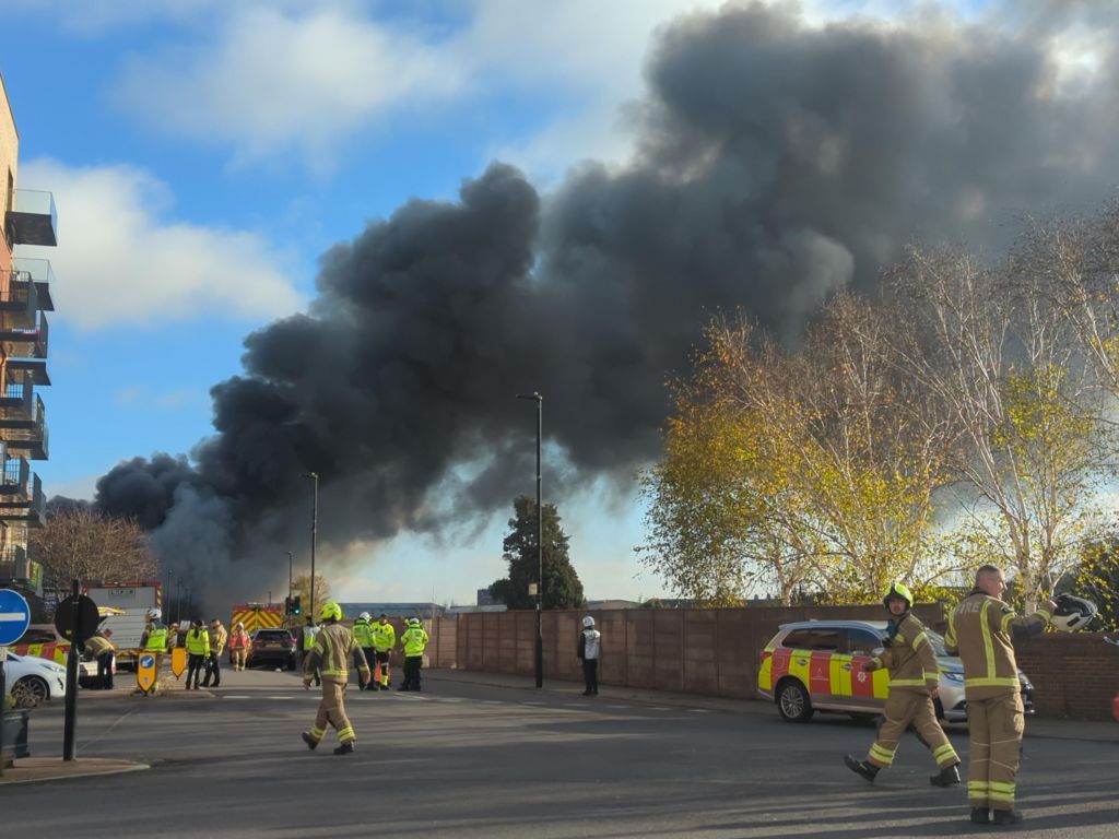 Large plume of black smoke emerges from a building (left)
In the foreground are firefighters in uniform including yellow helmets.