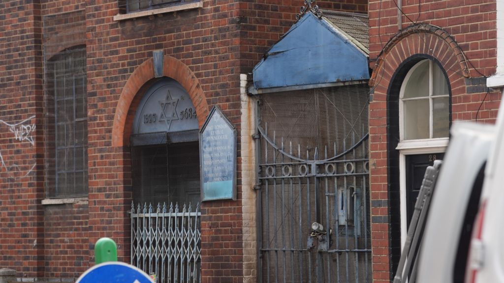 A close-up of a red brick building featuring an arched entrance with a Star of David and "1923" and "5684" inscribed above the door. A blue sign for the "East London Central Synagogue" is mounted on the wall. To the right of the entrance is a tall, padlocked metal gate topped with a weathered blue gabled roof.
