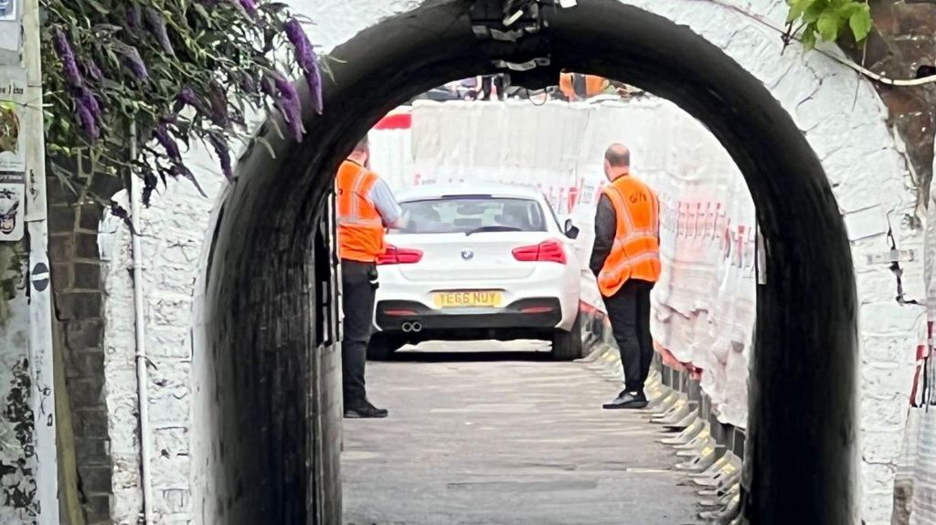 A narrow walled footpath passing under a bridge, with a white BMW car stopped in the middle and two men in orange high vis jackets looking at it