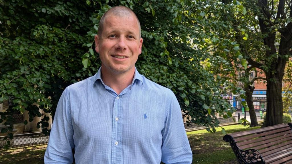 Mortgage adviser Ben Perks smiling, wearing a light blue shirt and standing in front of a bench and trees