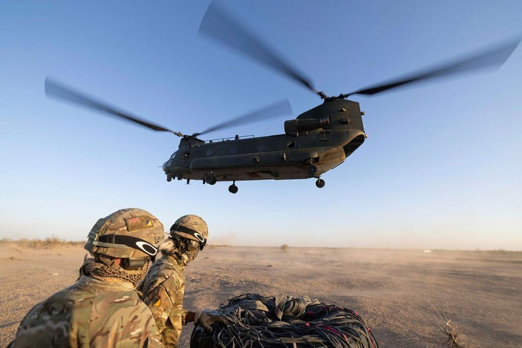Two joint helicopter support squadron soldiers look away from camera while wearing camo gear and helmets, with a CH-47 Chinook mid air above them in a desert type environment. Image taken near El Centro USA in October.