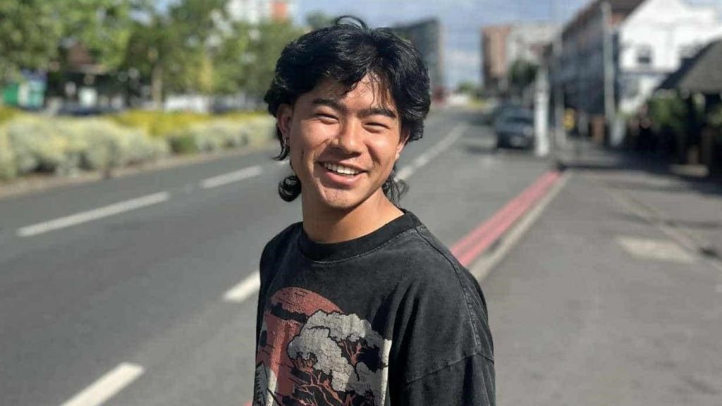 Yubin Tamang wearing a black t-shirt with a pattern stands on the side of the road and smiles. He has dark hair.
