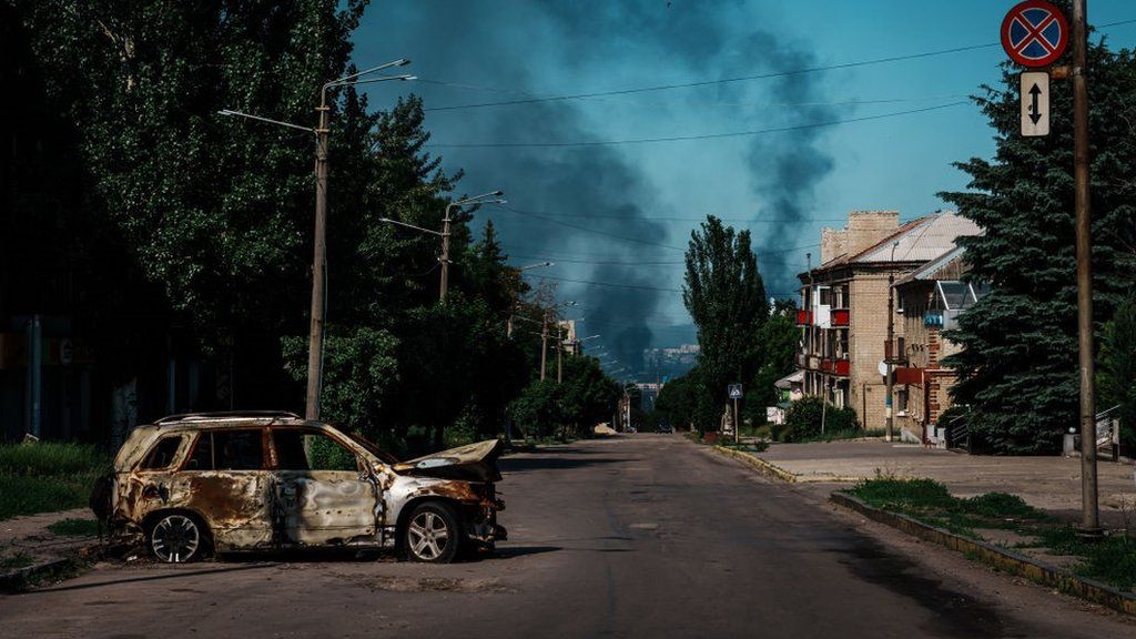 A destroyed car with smoke rising over Severodonetsk