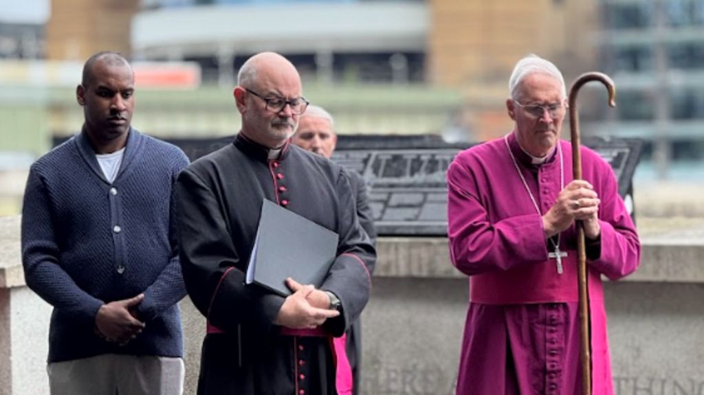 A number of people stand on a the walkway. One wears a bright pink ceremonial robe and holds a staff, while others in dark formal outfits stand nearby in a moment of reflection. 