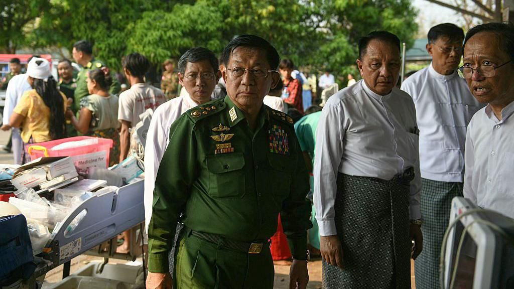 The military chief, dressed in army greens, looking around the compound, surrounded by other men.