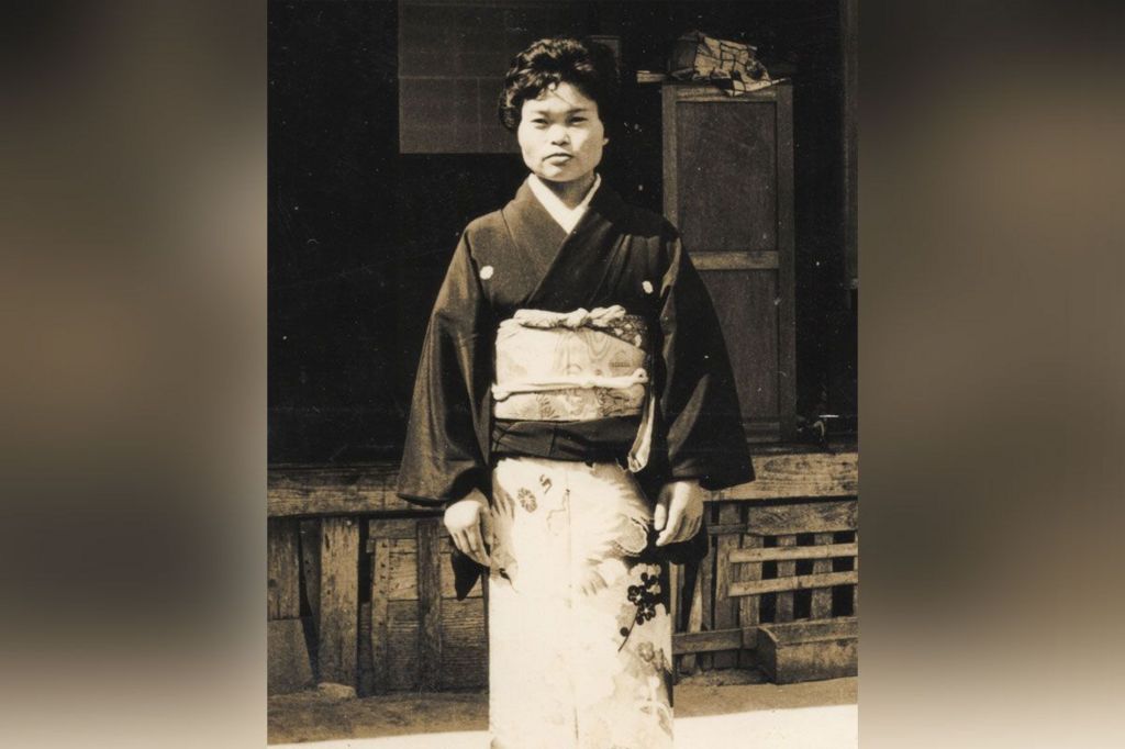 Black and white full length photo of Kiyomi Iguro. She has short dark hair and is wearing a traditional Japanese dress, of a black long sleeved top, and a white patterned wrap dress that stops at her feet.