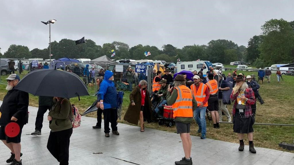 A group of festival goers behind a gate, one has their umbrella up.