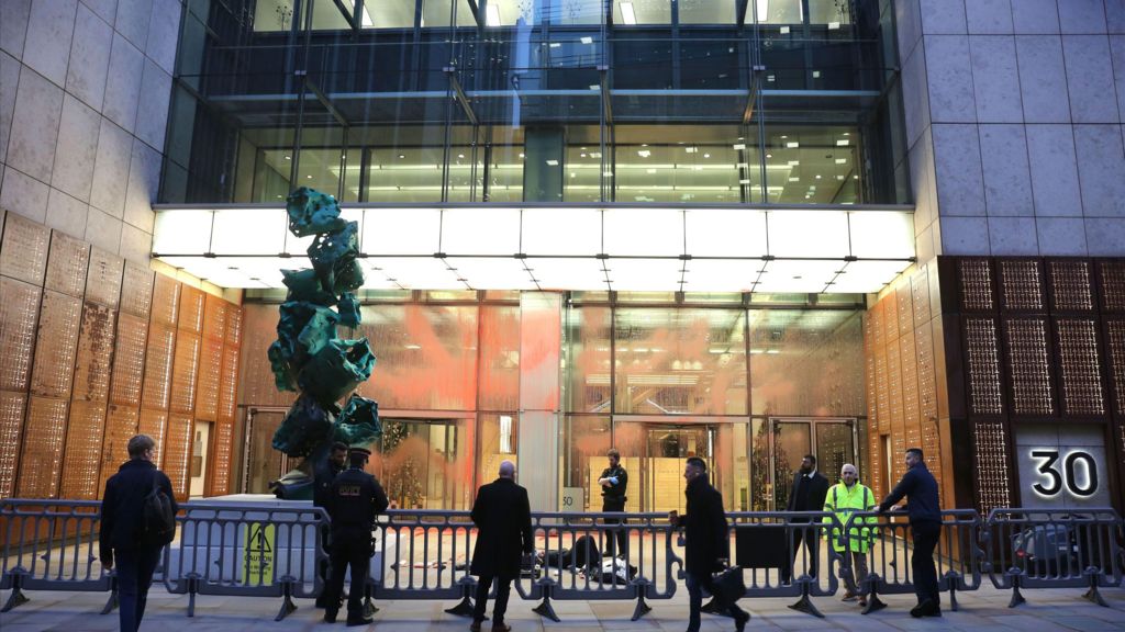 Police officers and security staff standing behind barriers outside an office building on Fenchurch Street, with red paint visible on the glass frontage following an early-morning protest.