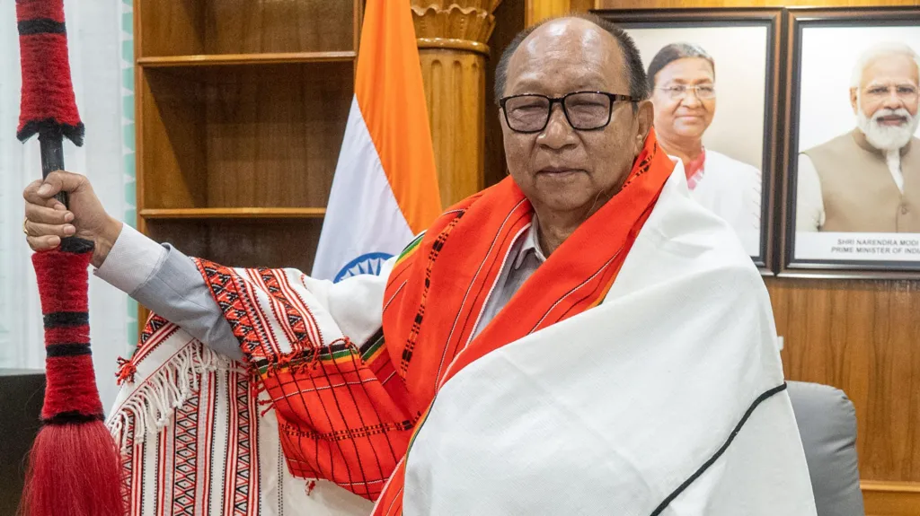 Yumnam Khemchand Singh wearing glasses, and a traditional white Naga shawl with red embroidery, as he stands in his office, after meeting a delegation of the Naga tribe last week. He holds a traditional Naga spear that has dyed-red tufts. Behind him there is an Indian flag and there are portraits of India's President Droupadi Murmu and Prime Minister Narendra Modi hanging on a wall. 