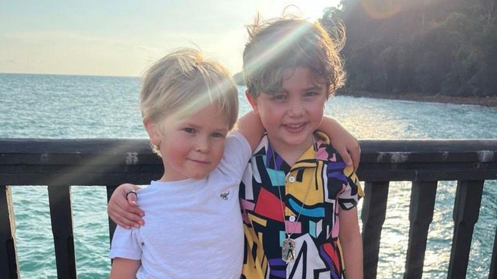 Fred and Beau stand on a balcony in front of the sea. They have an arm wrapped around each other. Fred has blonde hair and wears a white T-shirt. Beau has curly brown hair and wears a multi-coloured shirt. 