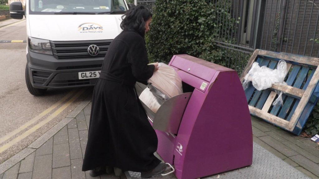 A young woman with black hair and wearing a black coat puts a bag of rubbish into a recycling chute in Tower Hamlets