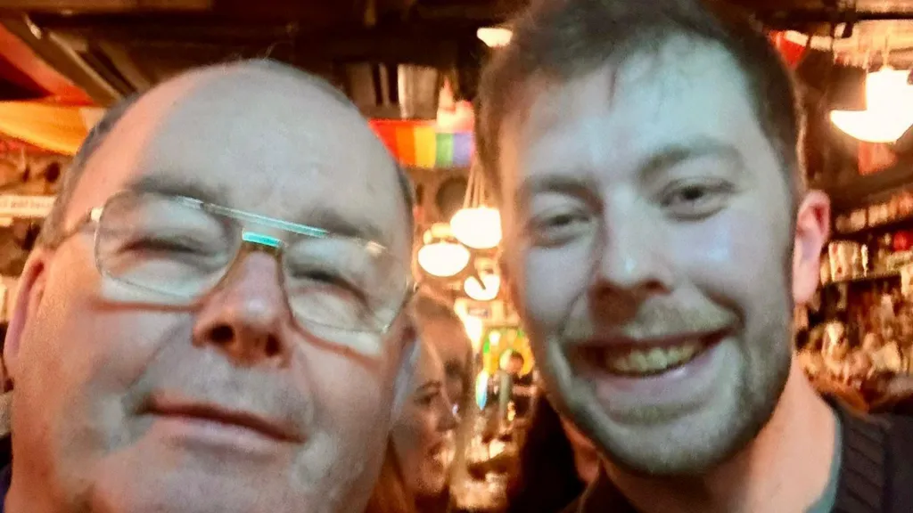 Two men appear close to camera in a selfie shot. The man on the leftis bald and wear glasses,  while the man on the right has short brown hair and is smiling. They are standing in a pub.