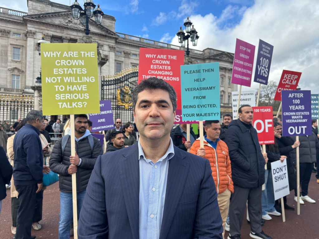 A man smiling for a photo. He is dressed in a suit standing in front of the protest placards outside the palace.