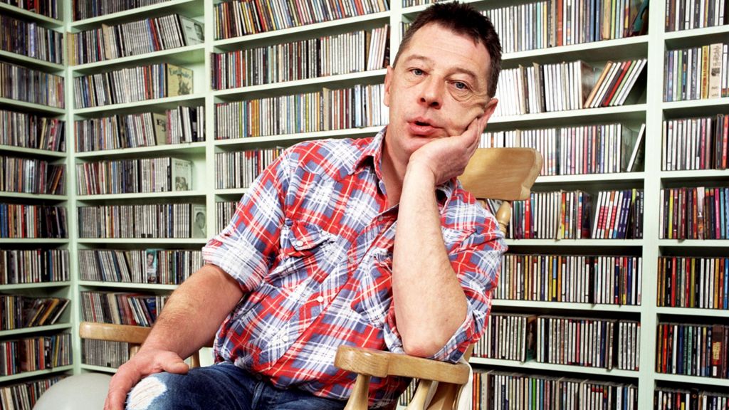 Andy Kershaw in a checkered shirt looking at the camera while in front of his record collection
