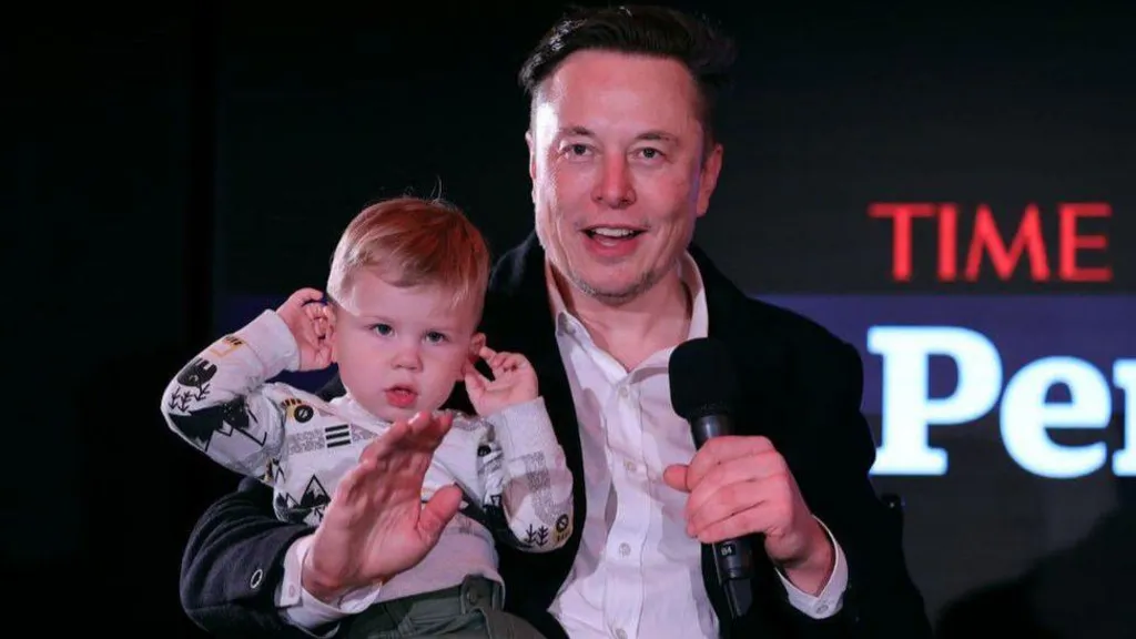 Elon Musk speaks on-stage at the TIME Person of the Year event in December 2021, while holding his son - who has his hands raised up to his ears.