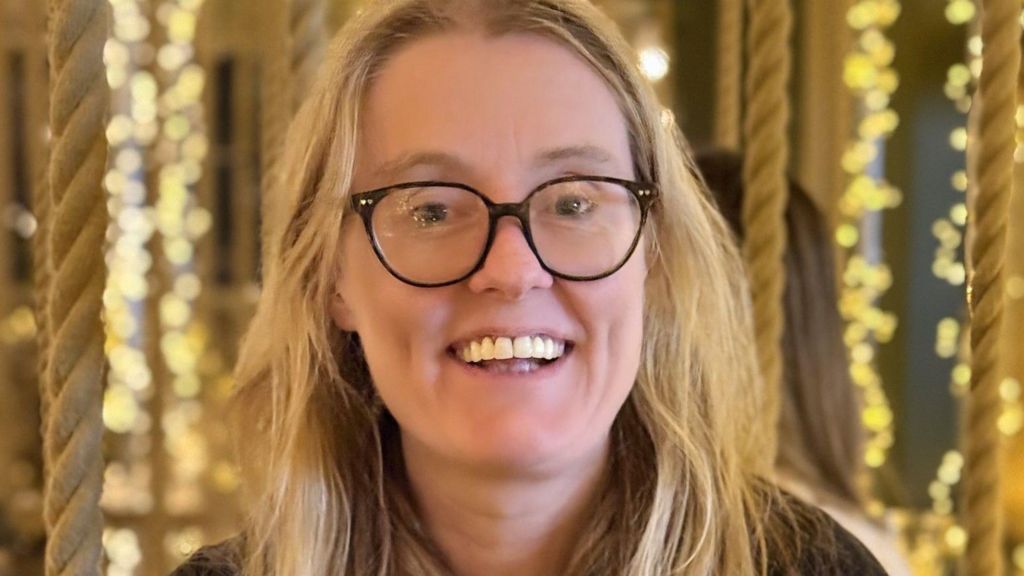 Head and shoulders image of Ellen Mulvey with long blond hair wearing black glasses and a brown top. She is sitting on a swing style seat held up with ropes in a restaurant. There are fairy lights in the background. She is smiling.
