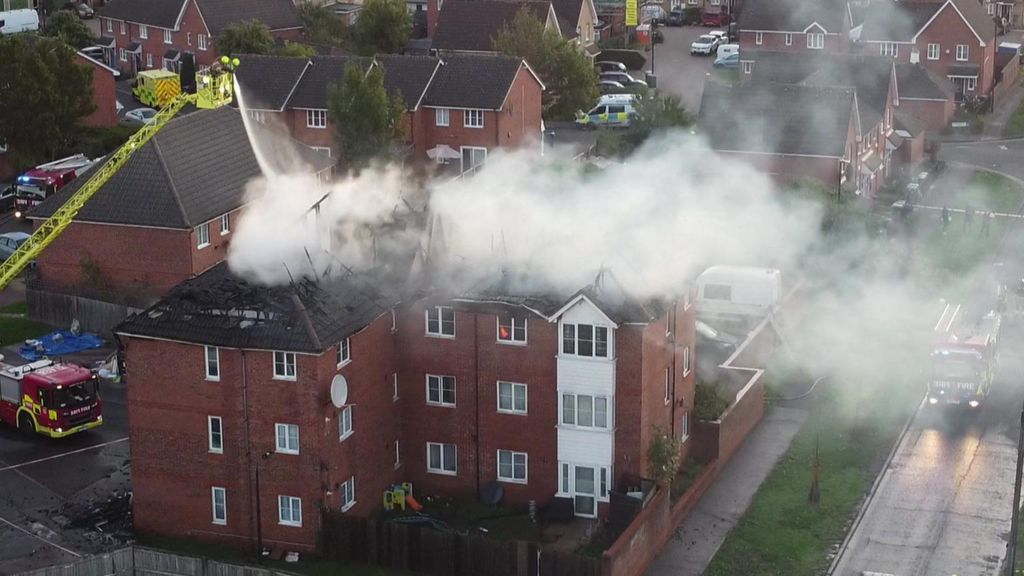 Aerial view of firefighters tackling a blaze in a residential area. Smoke billows heavily from the roof of a three-storey red-brick apartment block, which has been badly damaged, with its roof largely destroyed. Several fire engines and crews are visible, including one using an extended yellow ladder to spray water onto the fire. 