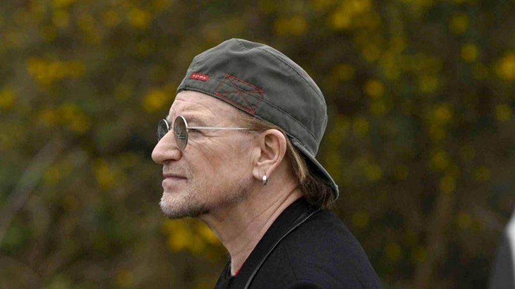 Shows Bono with baseball cap back to front and wearing glasses