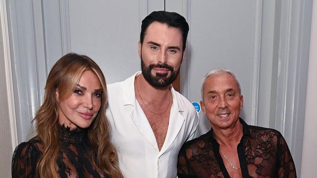 Lizzie Cundy, Rylan Clark and Bruno Tonioli pose in front of a white door. Lizzie is wearing a black lace outfit with long curled hair; Rylan, who is in the centre, has on a white shirt and thin chain; Bruno, on the right, has a black lace shirt and a long chain.