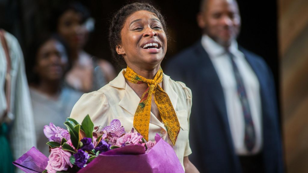 Cynthia Erivo smiles at the end of a performance of The Color Purple on Broadway in 2014. She is wearing a cream blouse, yellow neckerchief and is holding a purple bouquet.