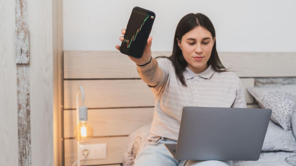Young woman sitting on a bed with a laptop on her legs and holding out a mobile phone with a graph on the screen.