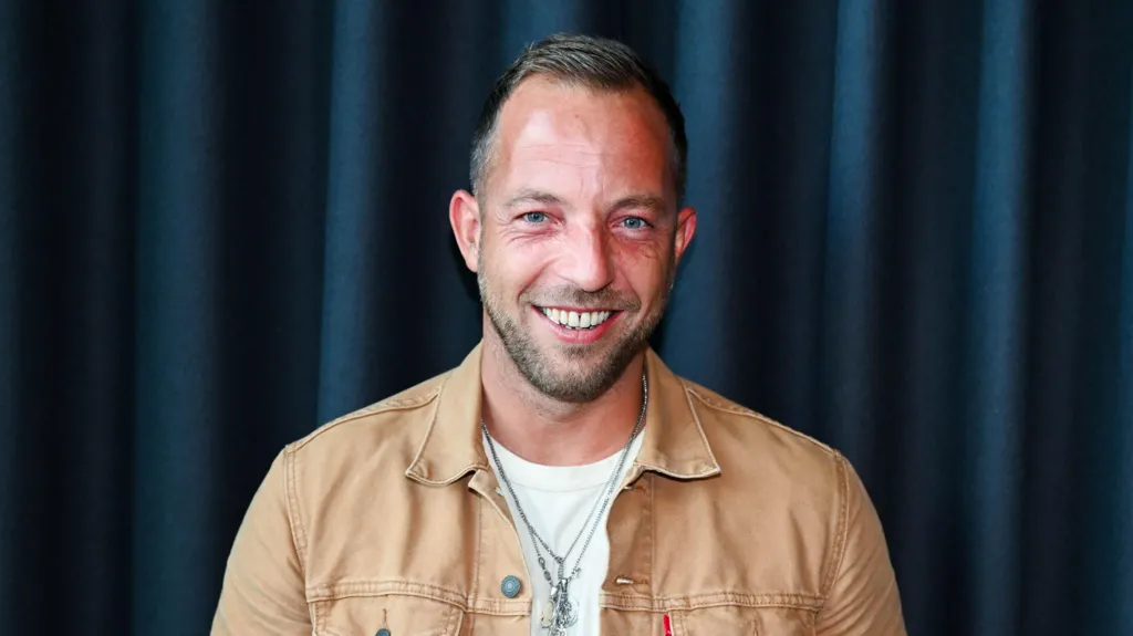 Musician James Morrison wearing a tan shirt and white t shirt. He has short dark hair and a short bead. 