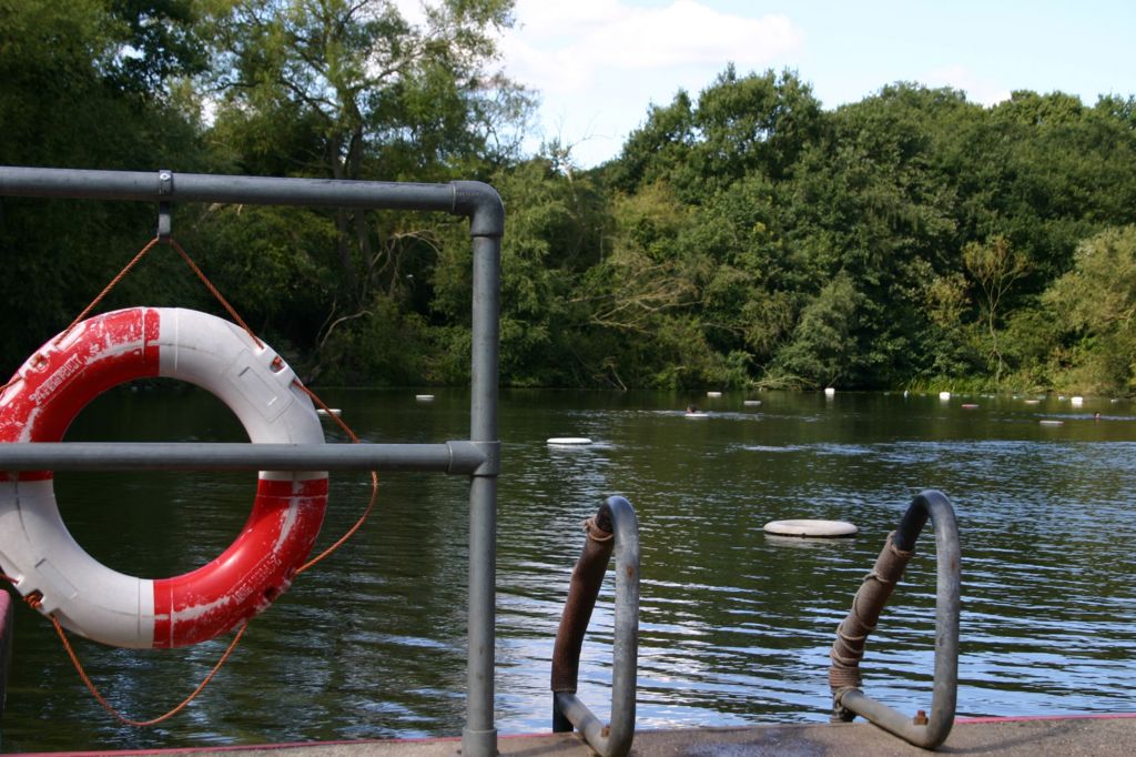 Life ring on a metal railing beside a leafy outdoor bathing pond with steps leading into the water at Hampstead Heath