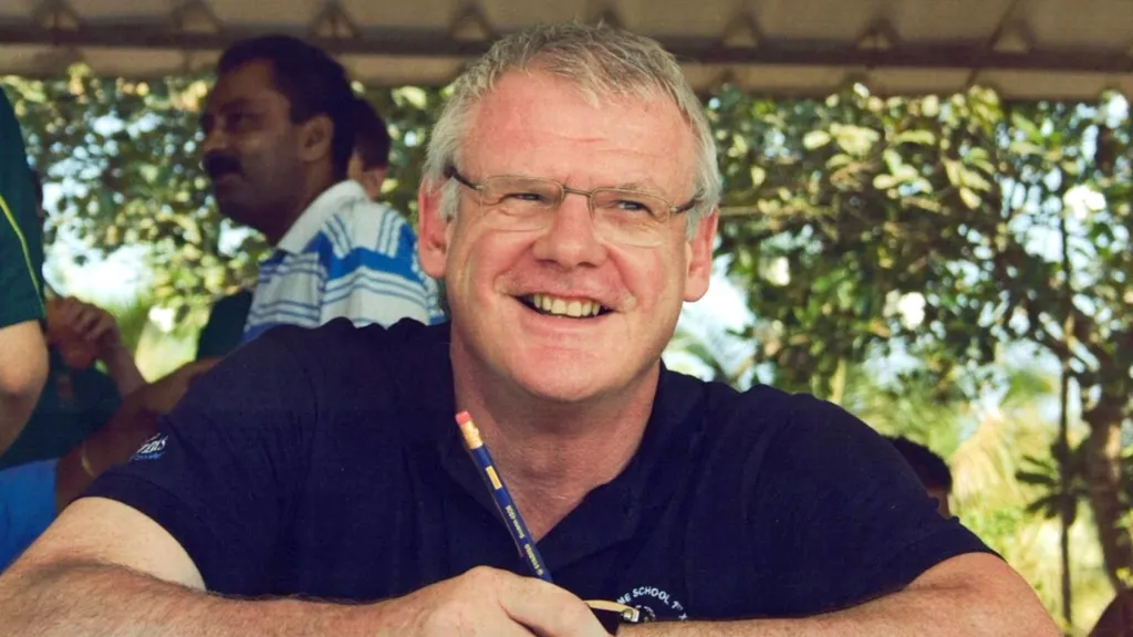 Andrew is wearing a blue polo shirt and spectacles. He in the shade, but it is a sunny day behind him. There are a few people visible in the background on the left-hand side of the image.