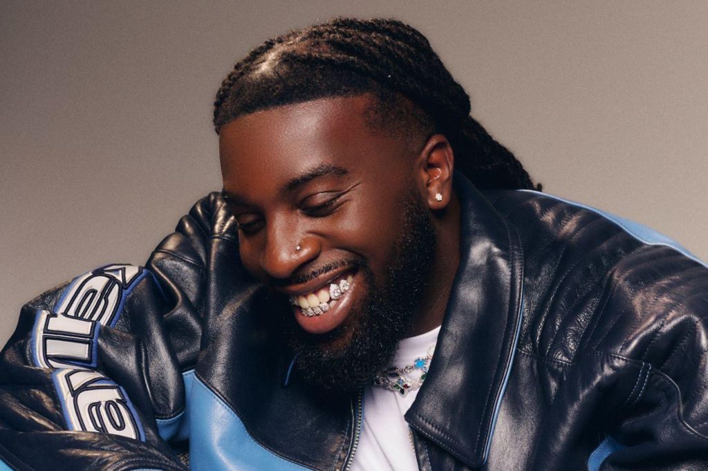 Mark Orabiyi looking down smiling. He is wearing a black and blue leather jacket over a white T-shirt