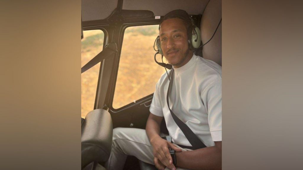 A man in a white shirt sits in a helicopter with a headset on. He smiles at the camera.