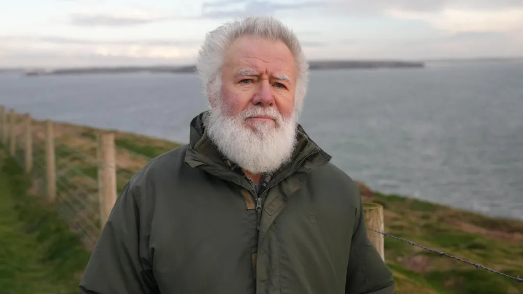 Retired BBC Wales reporter Hefin Wyn at St Anne's Head, Pembrokeshire.  He has white hair and a white beard and is wearing a green, waterproof coat.  Behind him can be seen the entrance to the Milford Haven waterway.