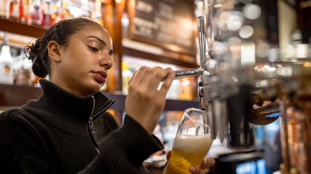 A lady pours a pint in a pub in London. She wears a black zip-up jacket, wearing lipstick and has her hair tied up.