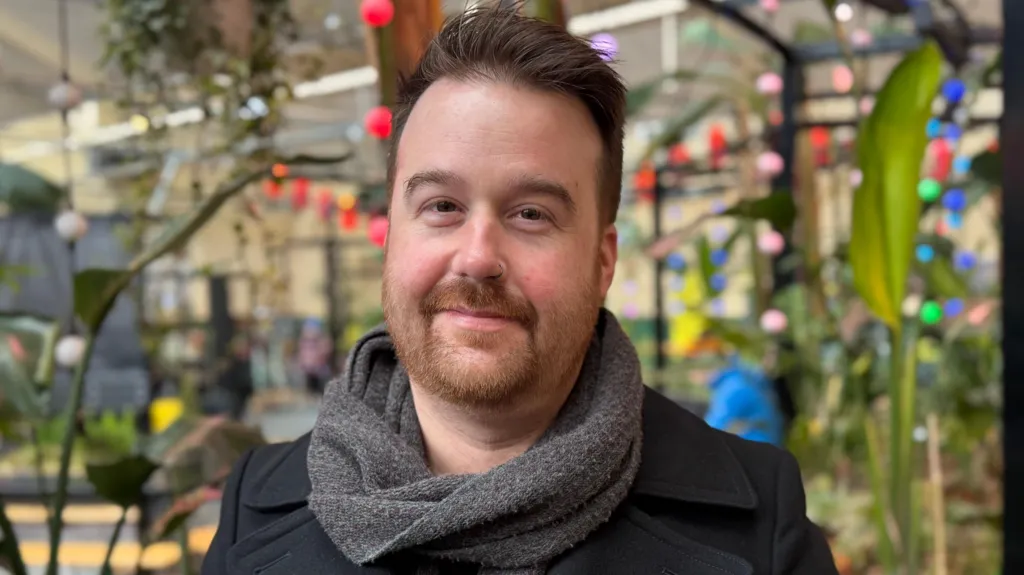 Michael is smiling at the camera. He is wearing a grey scarf and black coat. Behind him there are green plants and colourful string lights hanging from the roof. 