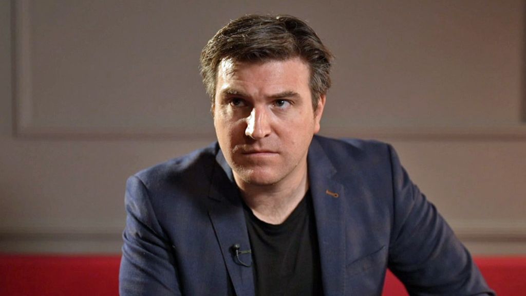 Headshot of Prof David Grimes who has short dark hair and is wearing a black t shirt and navy suit jacket. He is sitting on a red sofa with white wood panelled walls behind him.