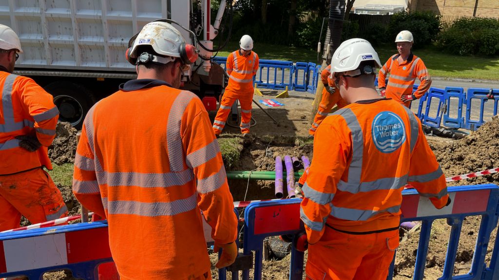 Engineers in orange Thames Water uniforms and white hard hats, examine damage to purple and green pipes lying in the ground