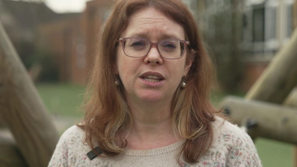 Linda wears a light coloured jumper, has shoulder length mid-brown hair, spectacles and a pearl earrings.