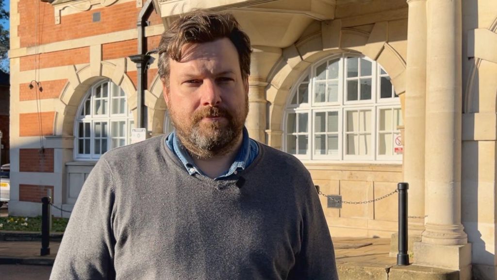 Man wearing a grey jumper, he has a light brown beard, short light brown hair, and behind him is a stone building with two arched windows, probably the Barnet Council building.