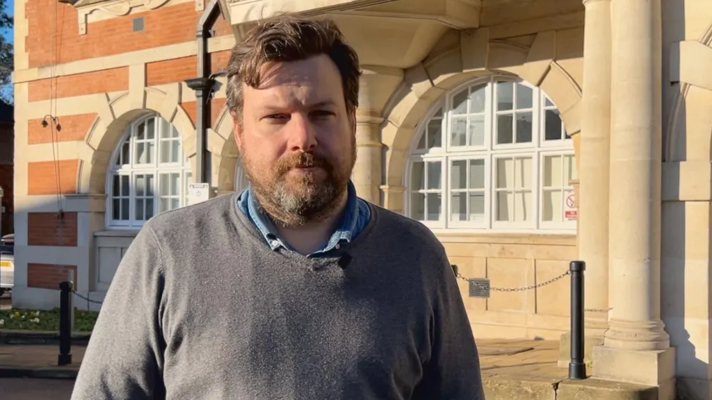 Man wearing a grey jumper, he has a light brown beard, short light brown hair, and behind him is a stone building with two arched windows, probably the Barnet Council building.