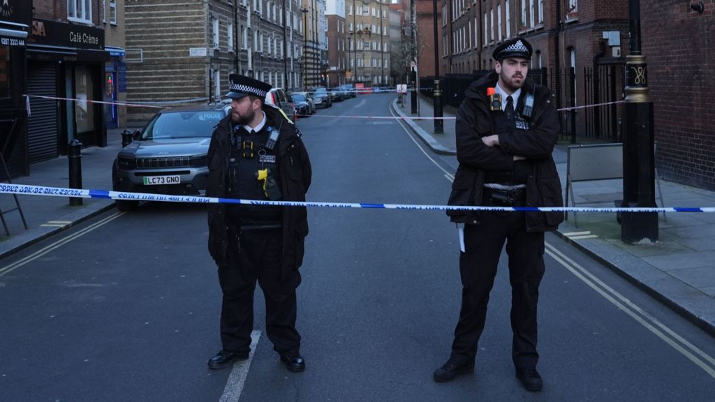 Two police officers standing behind police tape. Both are men dressed in black uniforms. Behind them is a street with high brick buildings either side and parked cars