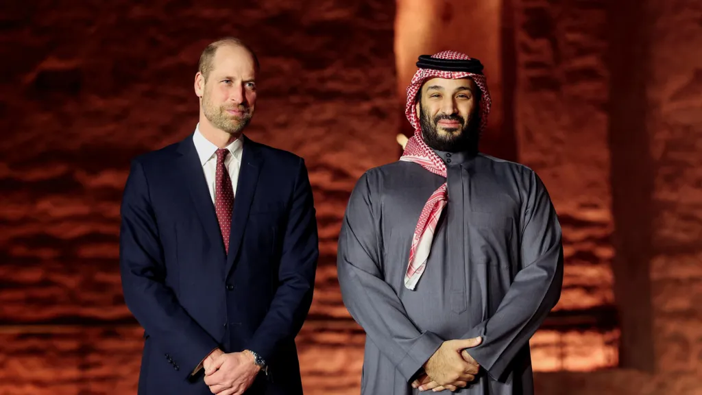 Prince William and Mohammed bin Salman pose for a photograph. Both men stand with their hands clasped in front of them.