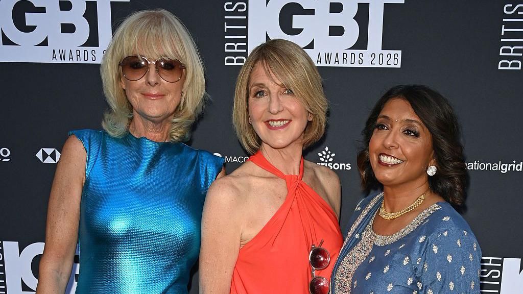 A photograph of Jane Moore, Kaye Adams and Sunetra Sarker from the waist up on the red carpet. The background is black with a logo reading 'British LGBT Awards'. Jane, on the left, wears a shiny blue dress and sunglasses with her hair in a blond bob. Kaye wears a red halter-neck dress with a similar hairstyle. Sunetra is on the right and wears a blue dress with gems, a gold necklace and silver earrings. She has dark hair.
