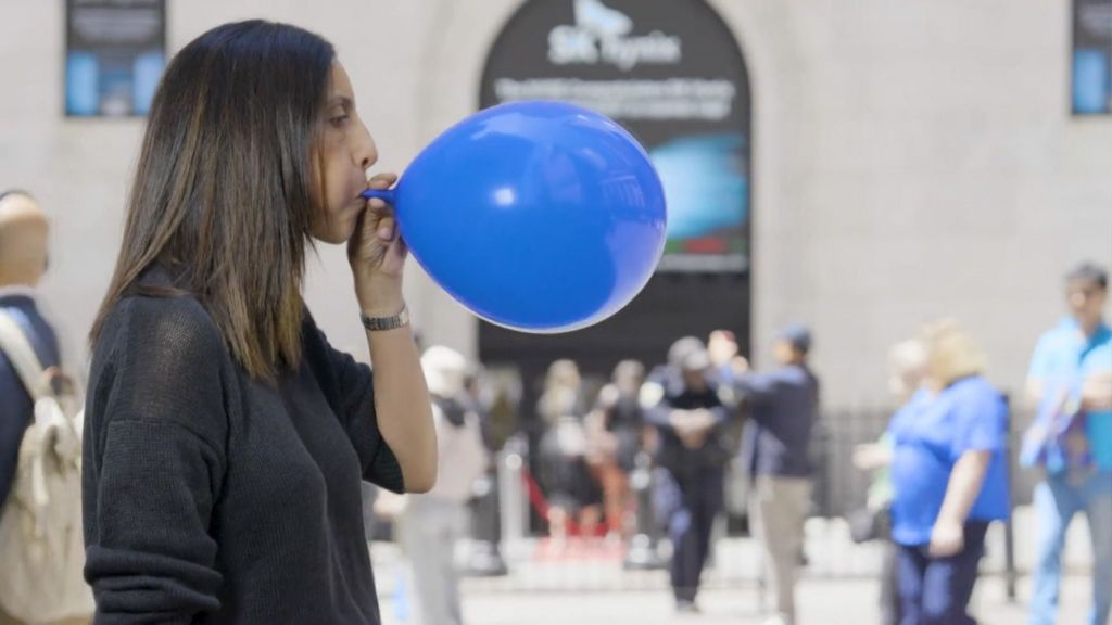 A woman stands in profile on a sunny day while blowing up a blue balloon.