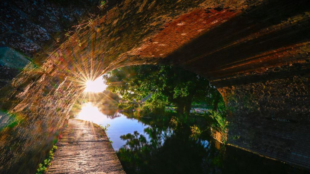 The sun peeks through under Nutshell Bridge on the canal in Stonehouse, near Stroud, Gloucestershire. It is reflected in the water