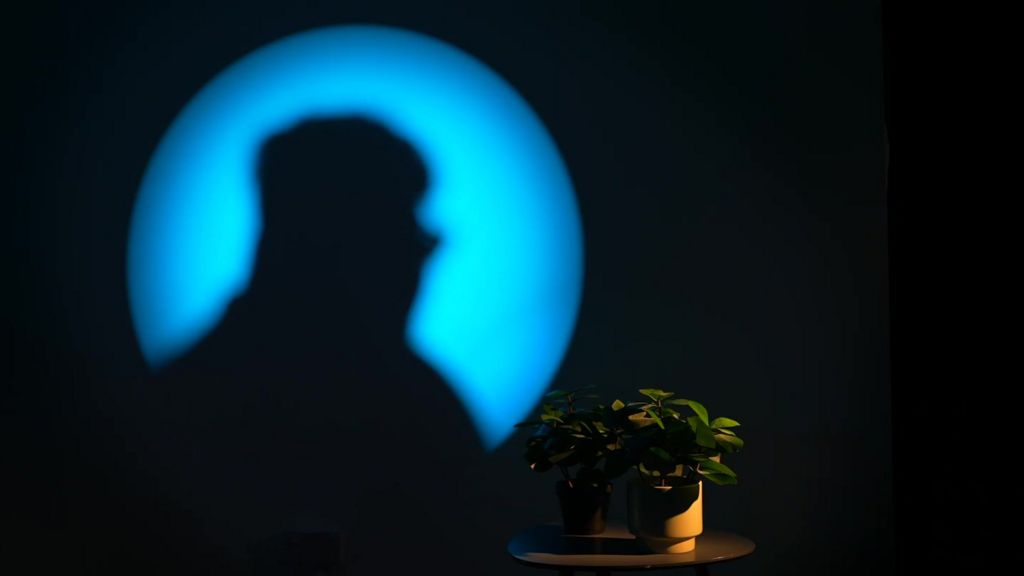 A silhouette of Tyler, filmed against a blue spotlight. They are sitting next to a table with a small pot plant on top