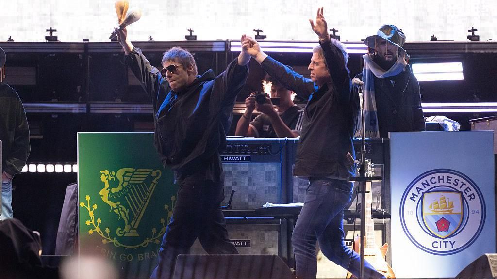 Liam and Noel Gallagher are raising their hands to the audience as they walk on stage during Oasis's reunion tour