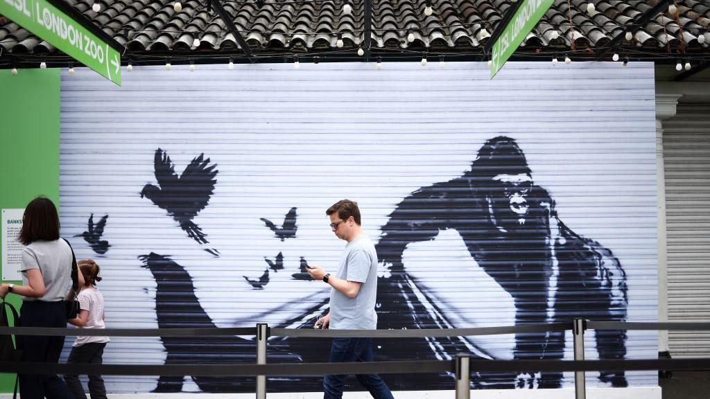 Visitors walk past a replica of the artwork by street artist Banksy, depicting a gorilla releasing animals, on the front of a shutter outside London zoo