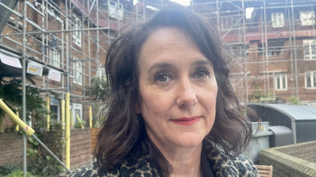 A middle-aged brunette woman wearing red lipstick and leopard-skin coat stands in front of a council housing covered in scaffolding