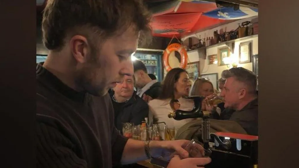 A man stands behind the bar in a packed pub as he poura pint. A number of patrons can be seen in the background of the image.
